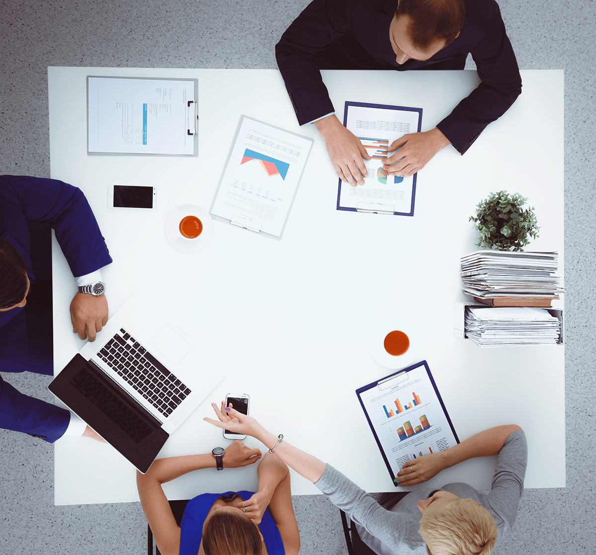 Un tavolo riunioni ripreso dall'alto: quattro persone al lavoro tra documenti, grafici stampati e un computer portatile.